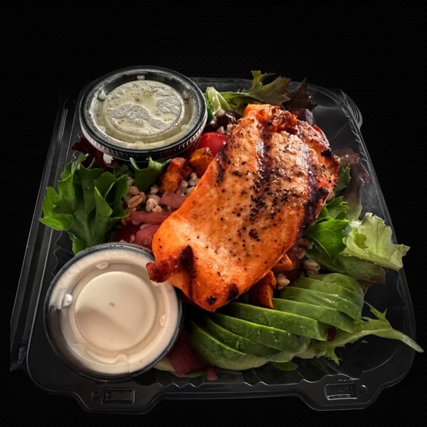 Grilled salmon salad with avocado, greens, and two dressing cups in a plastic takeout container.