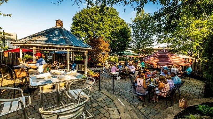 Patrons dine outdoors on a stone patio with a gazebo and large umbrellas under a sunny sky.