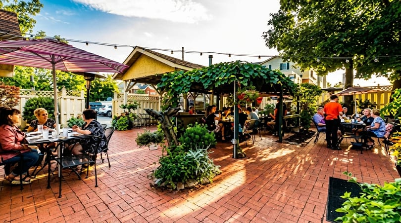 Patrons dine on a brick patio under umbrellas and a vine-covered pergola at a restaurant.
