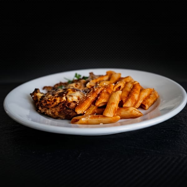 A white plate holds cheesy chicken parmesan and penne pasta with sauce and sesame seeds.