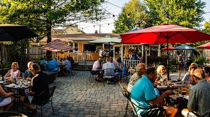 Patrons dine outdoors on a paved patio with umbrellas, string lights, and a distant bar area.