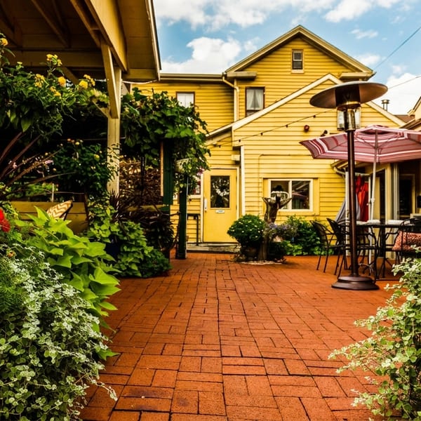 A charming outdoor patio with brick pavers, lush greenery, red flowers, and yellow buildings.