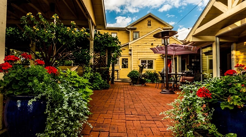 A charming outdoor patio with brick pavers, yellow buildings, and vibrant red and yellow flowers in planters.